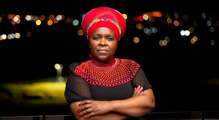 Ma Zwane in a red traditional Zulu headwrap and collar necklace, arms crossed against a night city backdrop